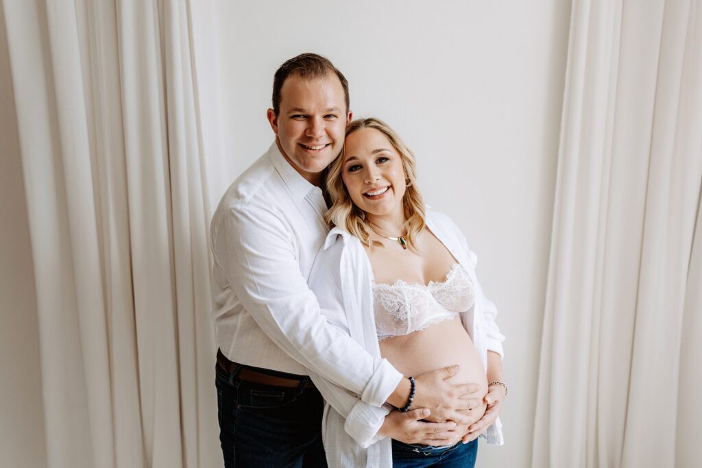 Pregnant couple smiling and holding belly during intimate boudoir-style maternity session in natural light Tampa studio