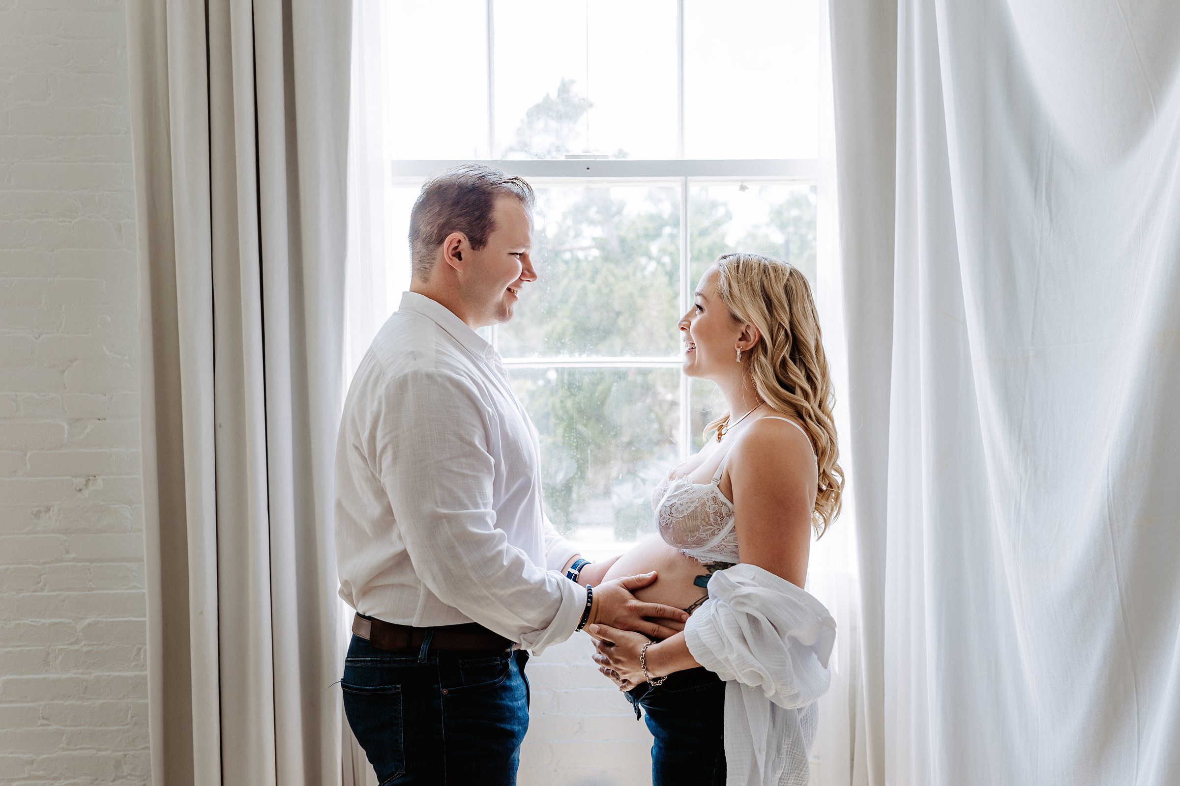 Couple standing by window, touching bellies and smiling during light-filled maternity session in Tampa Florida