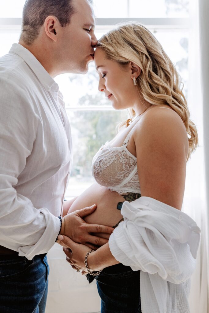 Close-up of couple holding pregnant belly, father kissing mother’s forehead during maternity photoshoot in Tampa studio