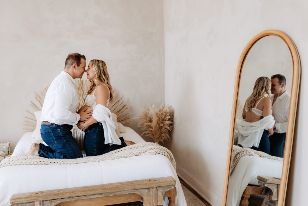 Romantic maternity portrait of couple leaning in for kiss on bed with mirror reflection in neutral Tampa photo studio