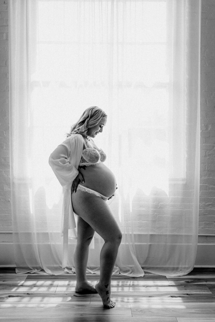 Black and white maternity photo of woman in lingerie cradling belly with backlight in soft natural light studio setting