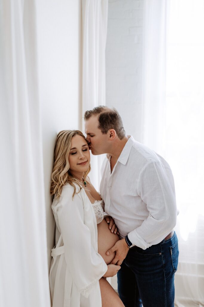 Partner kissing pregnant woman's forehead while holding her belly during intimate maternity portrait session in Tampa Florida