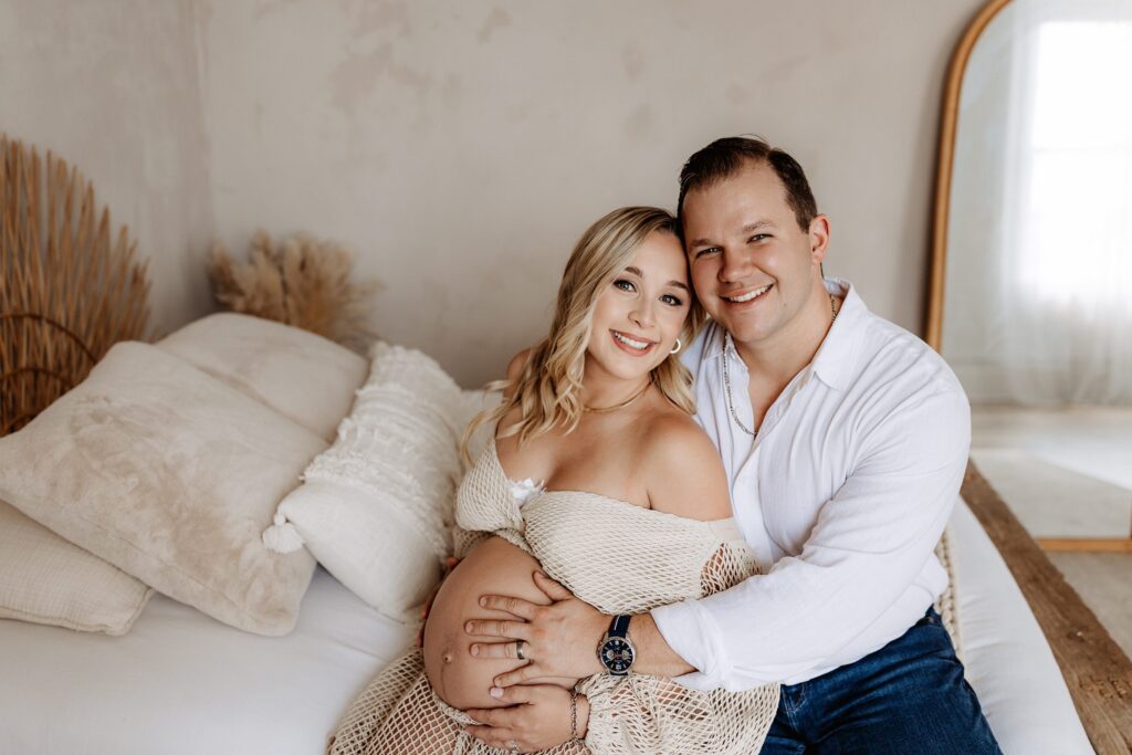 Maternity couple smiling together on bed, holding belly in soft and romantic Tampa lifestyle photo session