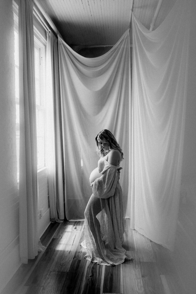 Black and white image of pregnant woman standing in window light with gauzy curtains during ethereal maternity photoshoot
