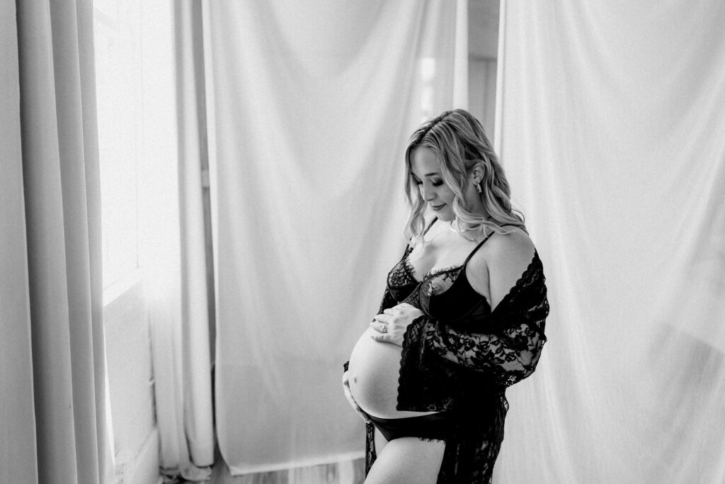 Black and white portrait of pregnant woman in black lace lingerie and robe, standing in soft backlight during moody boudoir maternity shoot