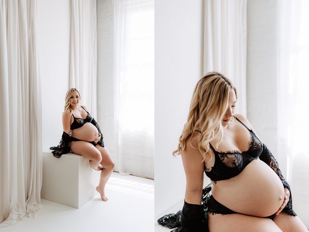 Maternity portraits of woman in black lace lingerie seated by white curtains, holding belly and smiling in elegant boudoir session