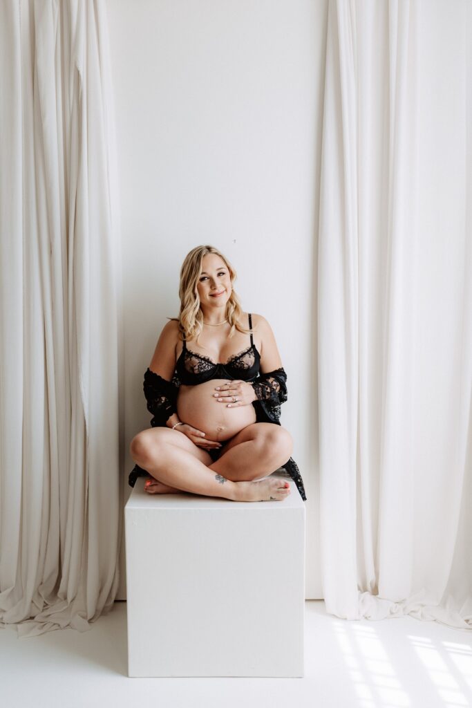 Pregnant woman sitting cross-legged on white cube in black lace lingerie, smiling softly in light-filled boudoir maternity studio