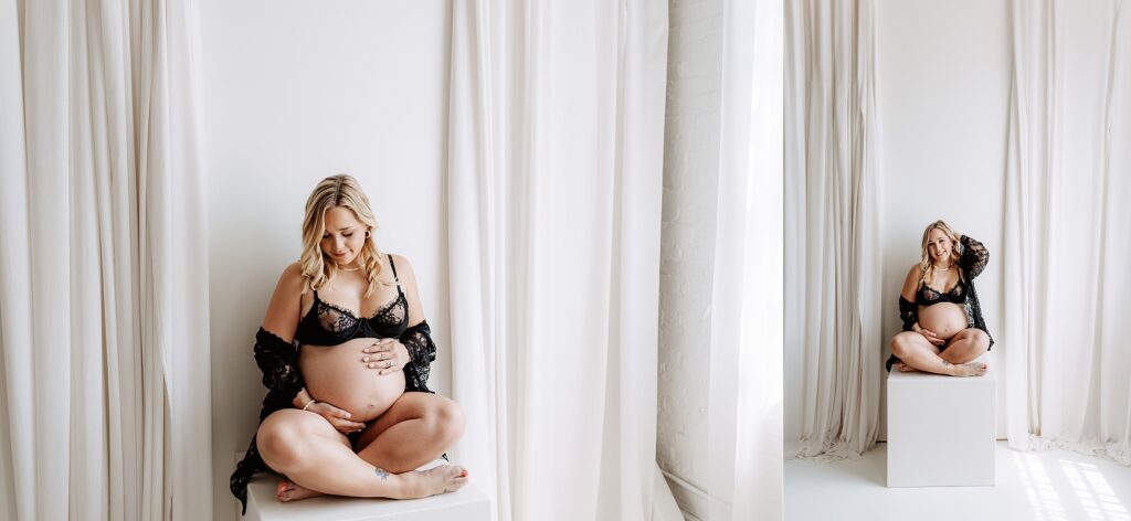 Pregnant woman in black lace lingerie sitting cross-legged on cube, holding belly and smiling during boudoir maternity shoot in Florida studio
