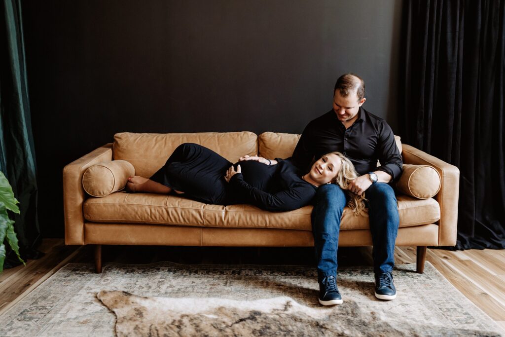 Maternity couple on camel leather sofa, woman in black dress lying across partner’s lap in cozy Tampa lifestyle session