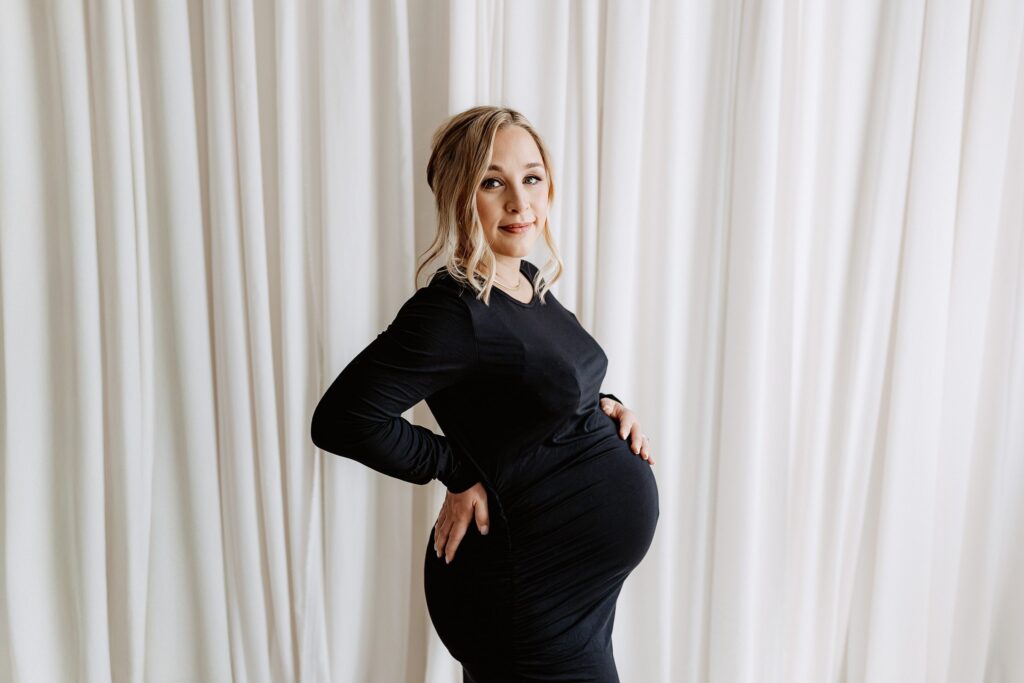 Confident pregnant woman in fitted black dress smiling with hand on hip, posed against white draped curtains