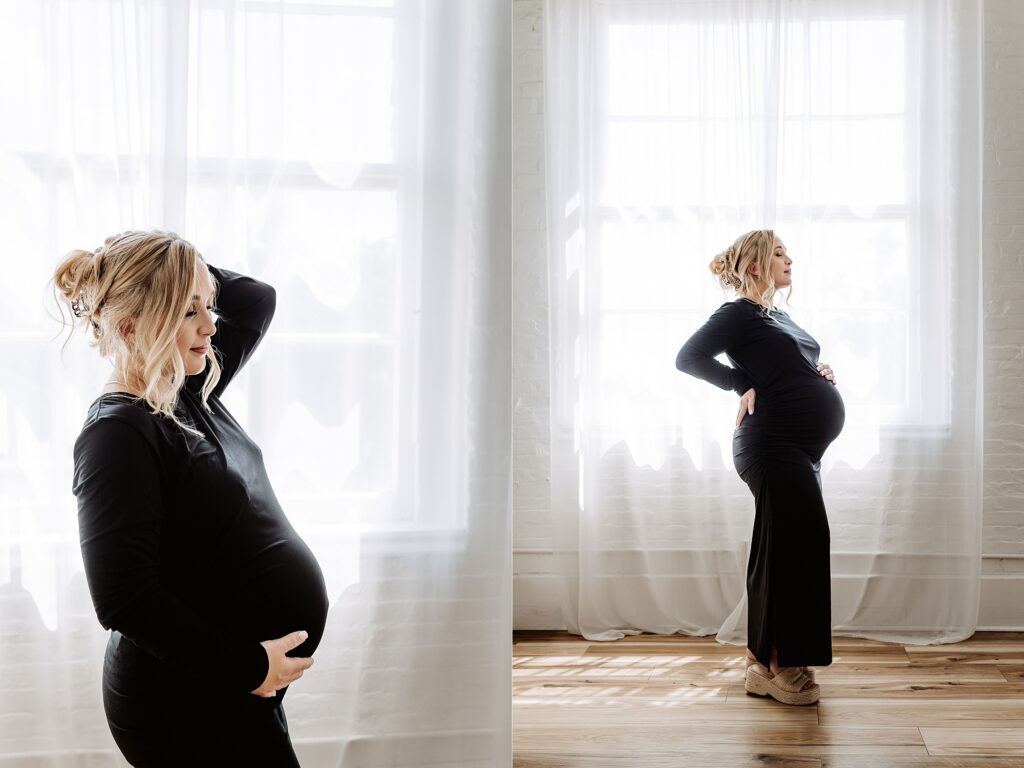 Pregnant woman standing by large window in black dress, one hand on belly, one in hair, during elegant maternity photoshoot in natural light studio