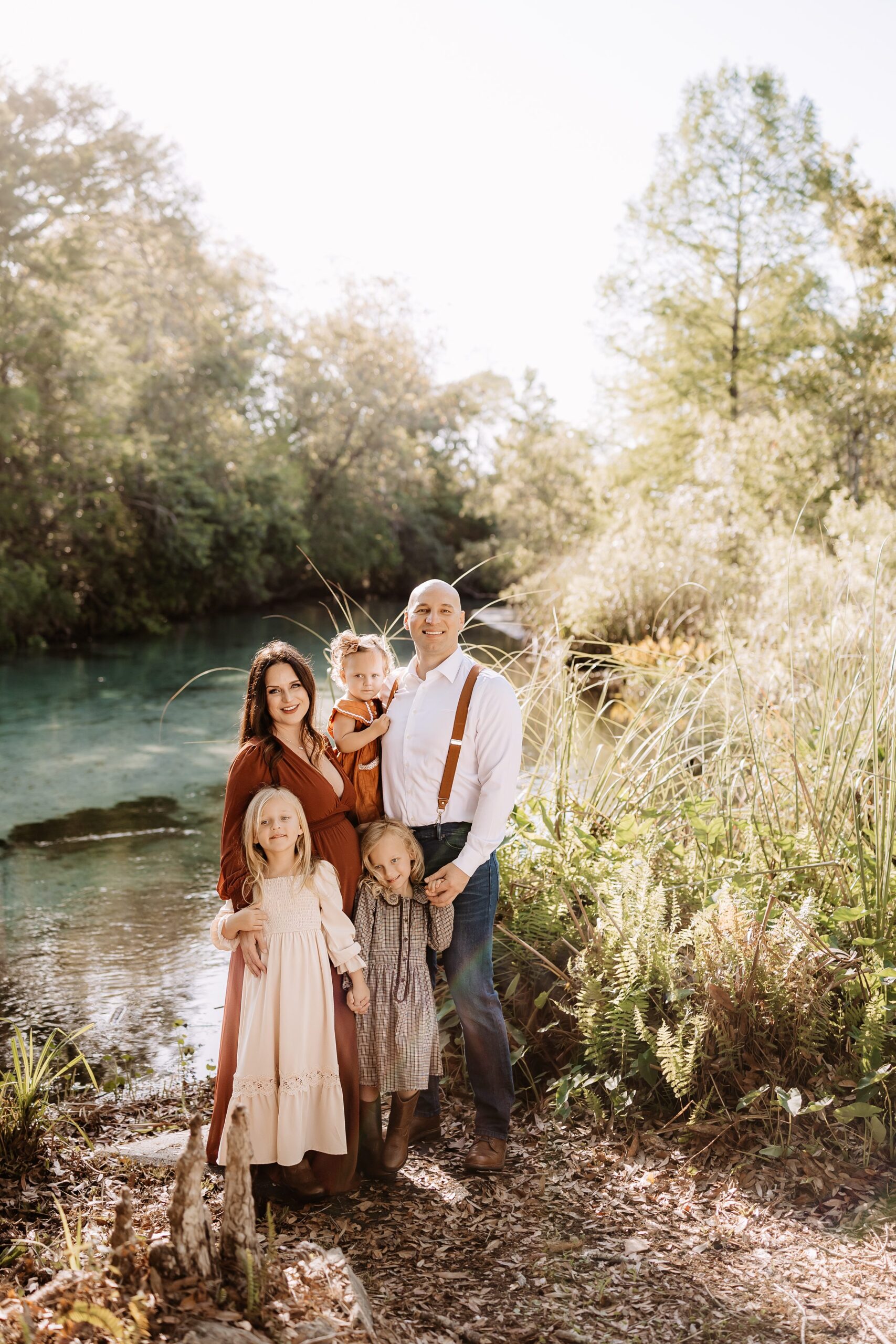 Pregnant mother and father with three young daughters posing beside clear spring water during outdoor family maternity photoshoot in Weeki Wachee, Florida