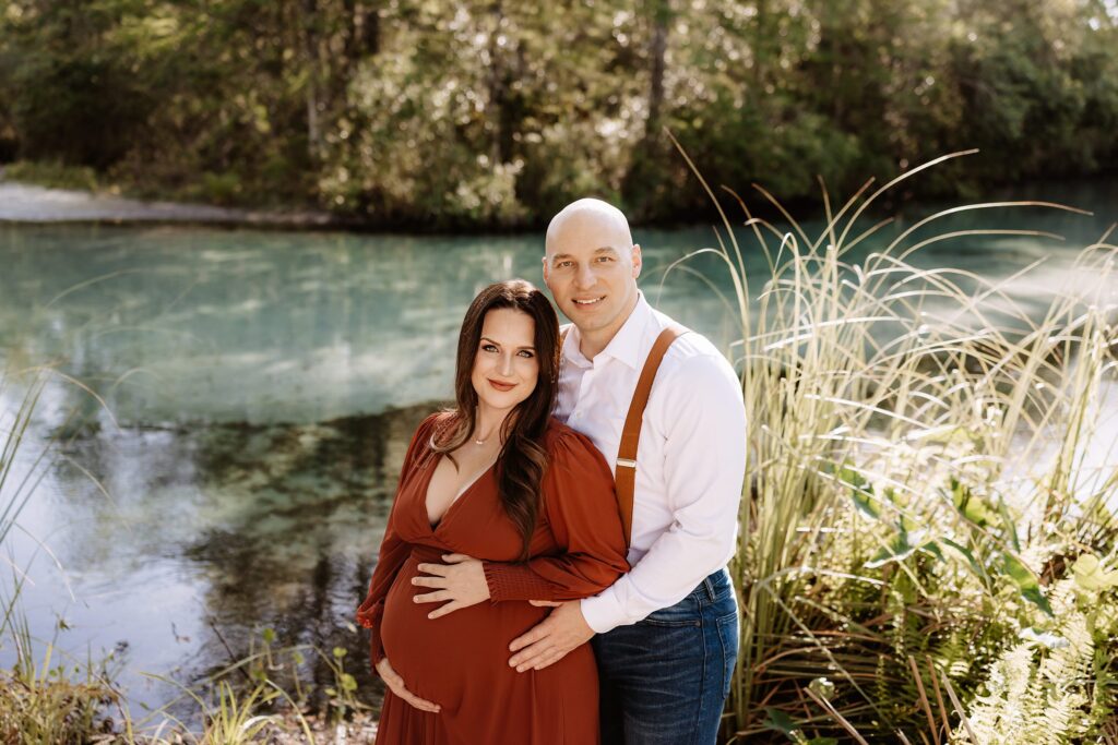 Married couple embracing with serene expressions, standing beside turquoise spring water during intimate maternity photos at Weeki Wachee