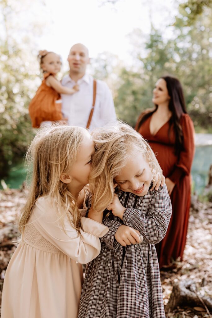 Two sisters laughing and hugging in foreground while parents watch lovingly in blurred background during playful family maternity session