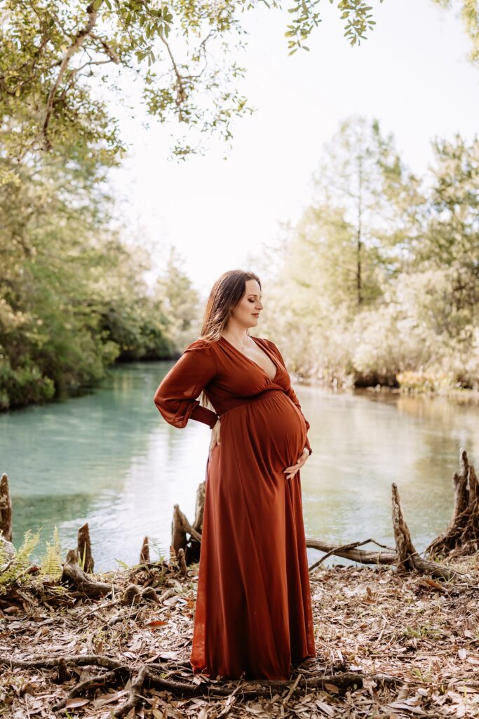 Pregnant woman standing confidently by cypress knees and riverbank in long rust dress during Florida outdoor maternity session