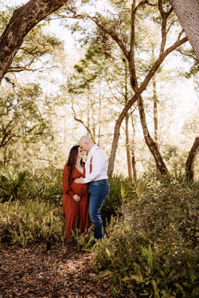 Pregnant woman and partner standing forehead-to-forehead among palmettos and oak trees during intimate forest maternity portrait