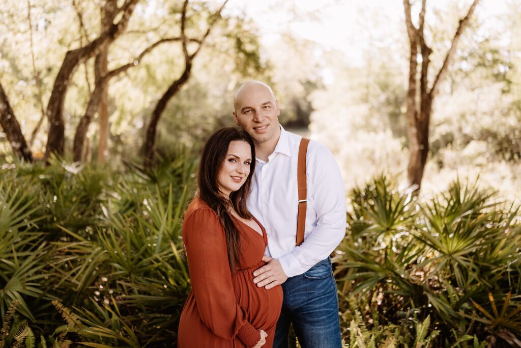 Expecting couple smiling at camera while standing close together in wooded Florida landscape, with sun filtering through trees