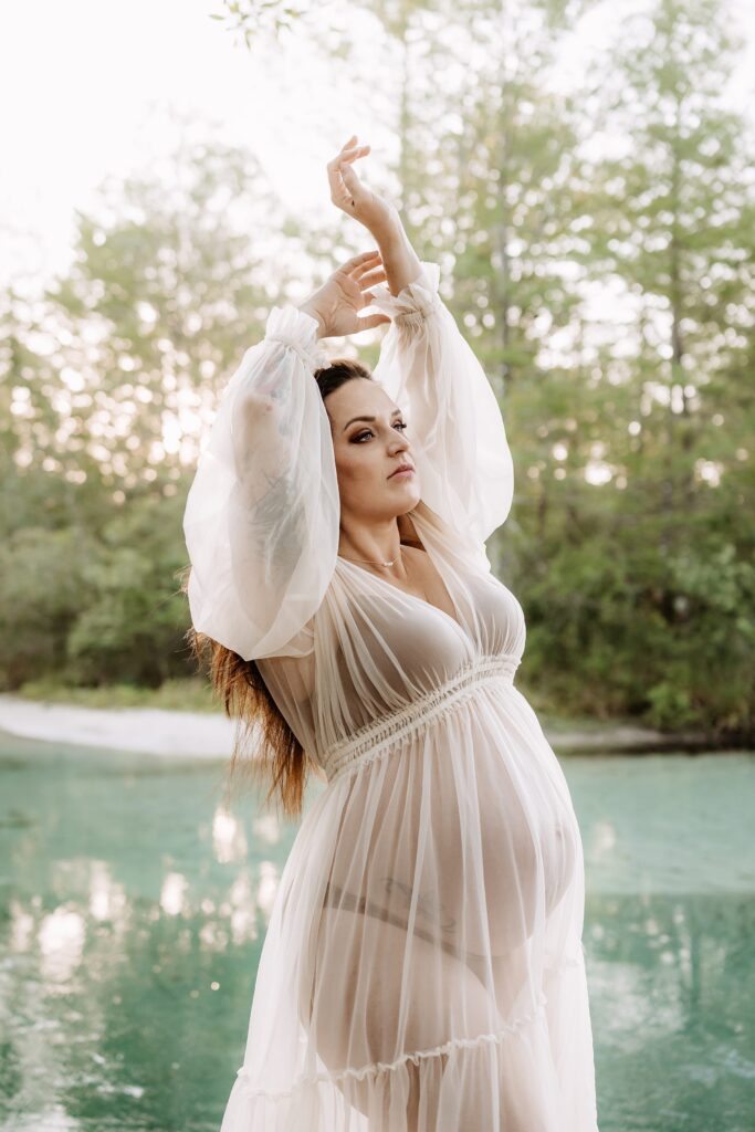 Maternity photo of woman in sheer white gown with arms raised gracefully, standing beside turquoise spring with dreamy forest in background