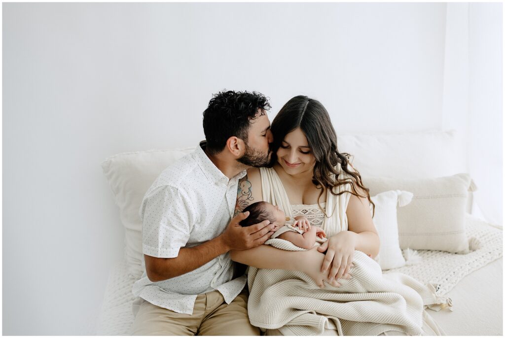 Picture of dad in white button up shirt kissing mom while holding newborn in Springhill Florida studio