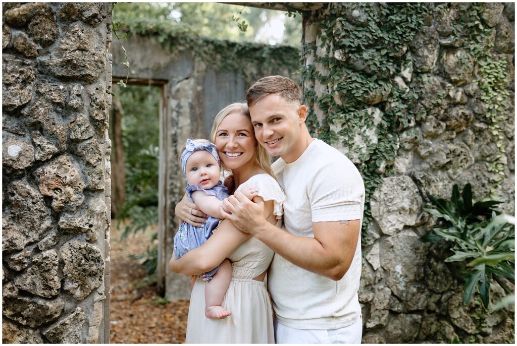 Baby in a blue and white floral dress being held by both mom and dad, creating a loving family moment.
