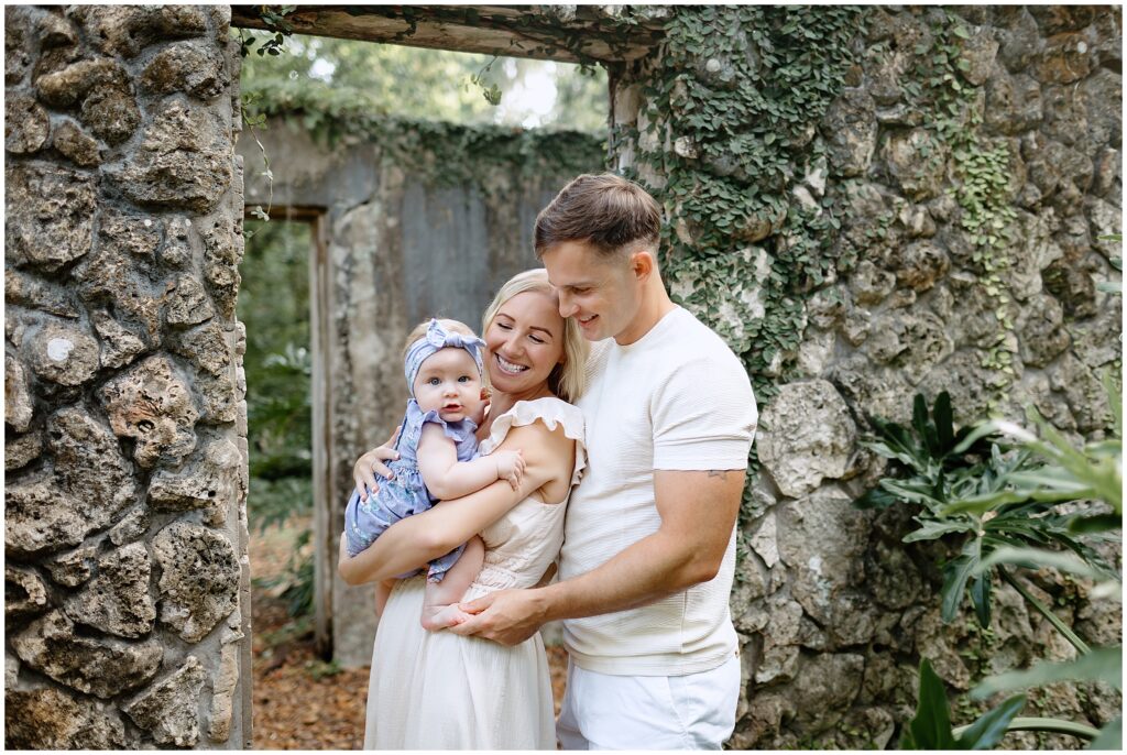 Baby in a blue and white floral dress being held by both mom and dad, creating a loving family moment.
