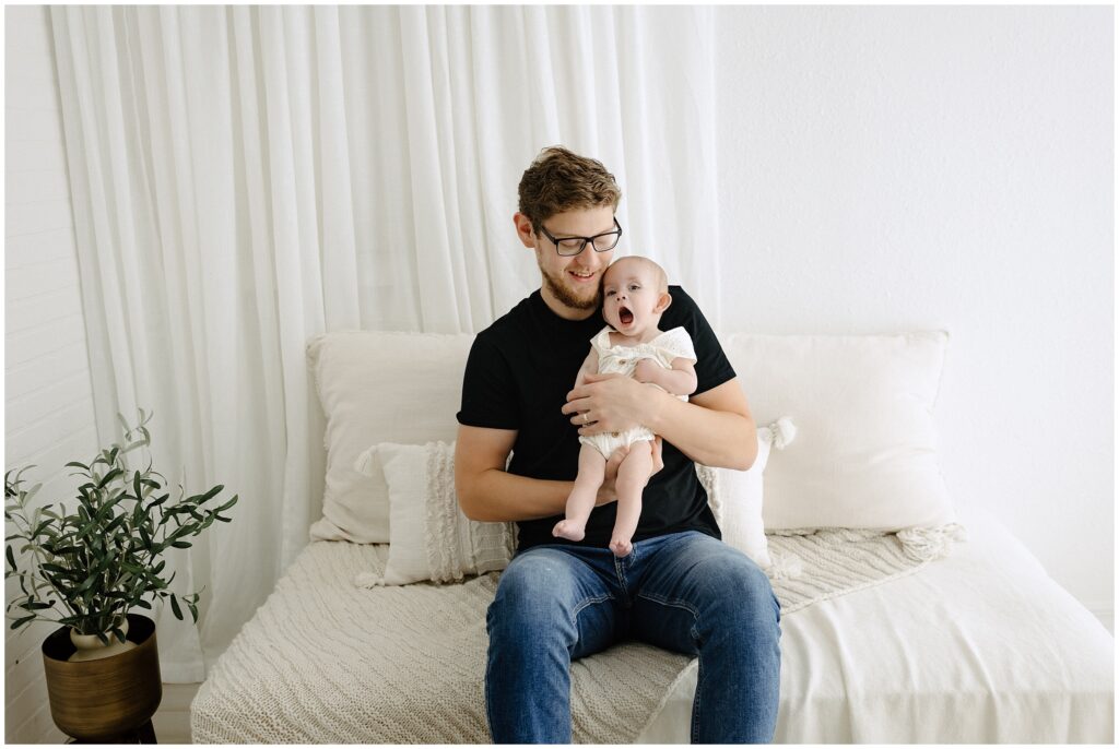 Yawning baby girl in dad's arms sitting on white bed Spring Hill Studio in Florida