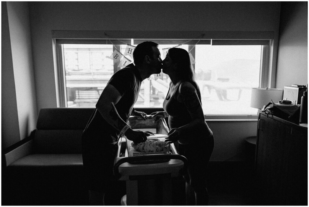 Black and white photo of mom and dad kissing in front of window with sleeping newborn baby, fresh 48 hospital newborn session in Florida