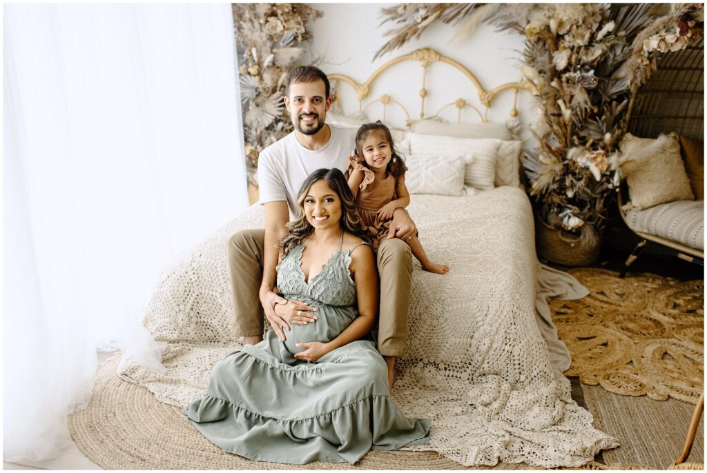 Beautiful family portraits of mom, dad, and daughter while holding pregnant belly on bed at Vibe Studio Spring Hill in Florida