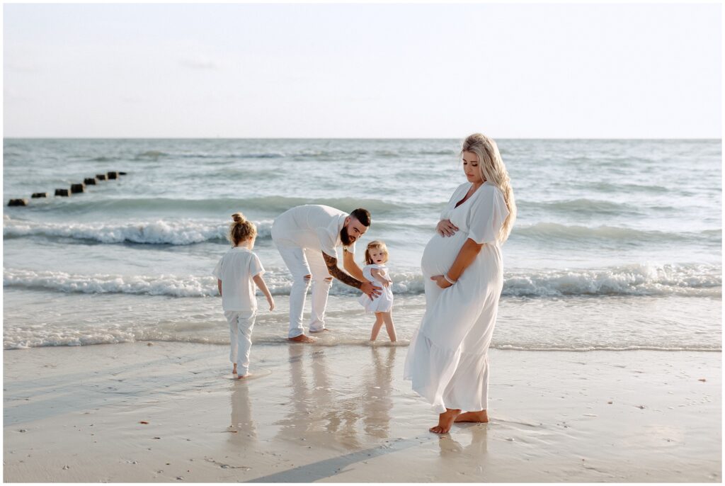 Mom holding belly during maternity session while dad and kids play in water in Tampa