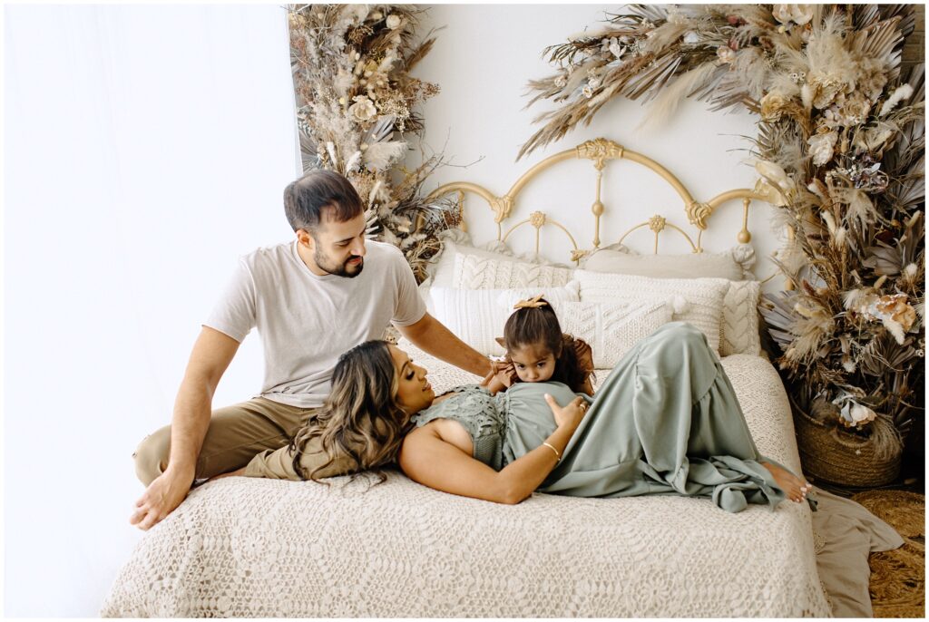 Little girl kissing mom's belly while she lays on bed in green dress with dad at Vibe Studio Spring Hill in Florida