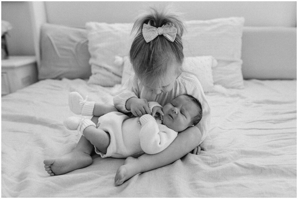 Black and white portrait of big sister holding baby brother on bed 