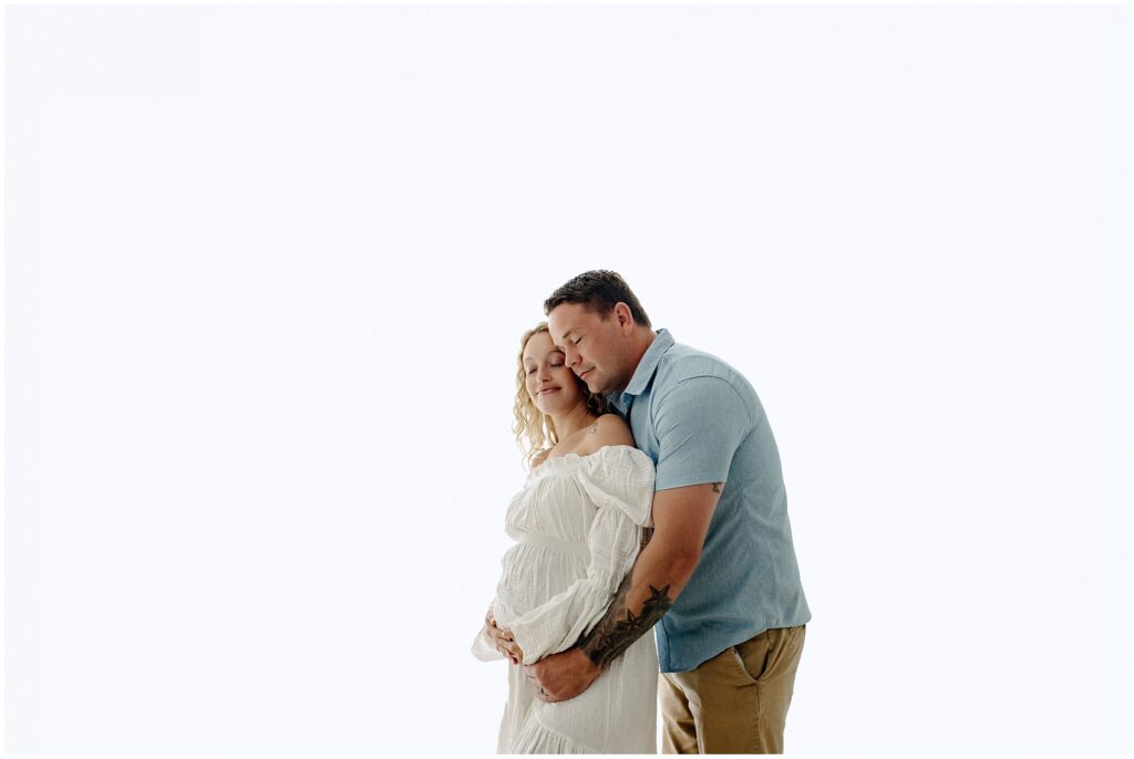 Pregnant couple smiling together in a white dress and blue polo at Spring Hill Studio in Florida