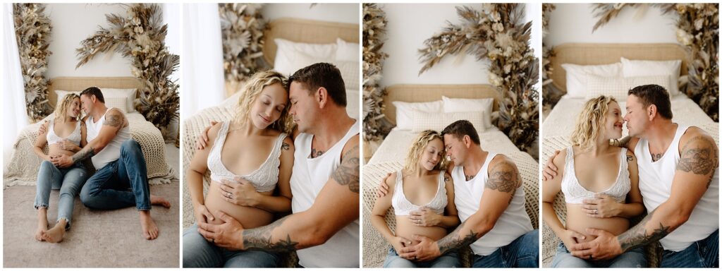 Pregnant woman with husband holding her belly, sitting in front of bed in white tops and jeans at Spring Hill Studio.
