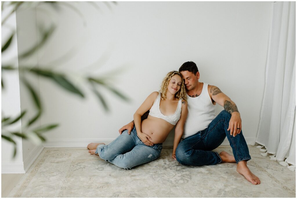 Pregnant couple in jeans and white tops, snuggling barefoot on the floor at Spring Hill Studio in Florida.
