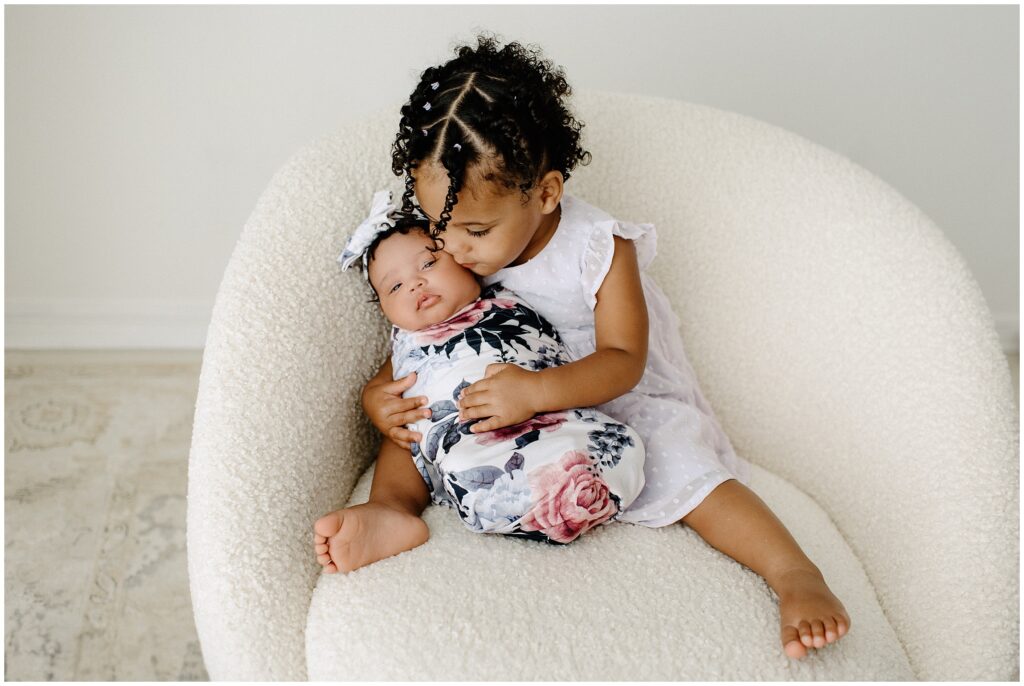 Newborn baby girl snuggling with big sister in swaddle in white chair newborn session at Spring Hill Studio in Florida