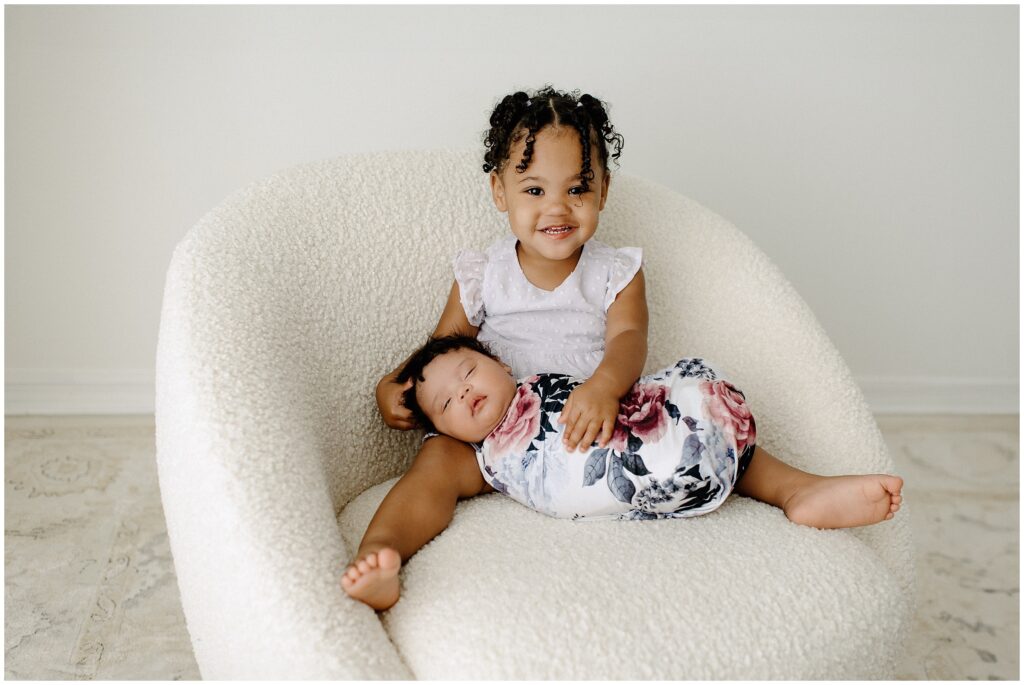 big sister cuddling with sleeping baby sister in white chair during newborn session at Spring Hill Studio in Florida